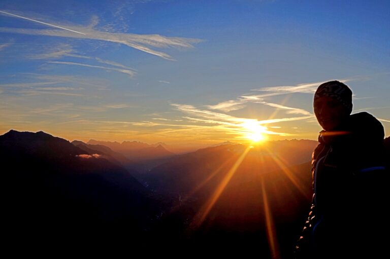 Eine Person steht als Silhouette bei Sonnenuntergang auf einem Berg, während die Sonne ihre Strahlen auf die entfernten Gipfel wirft. Der Himmel ist in Orange- und Blautönen gehalten und schafft eine ruhige und dramatische Landschaft.