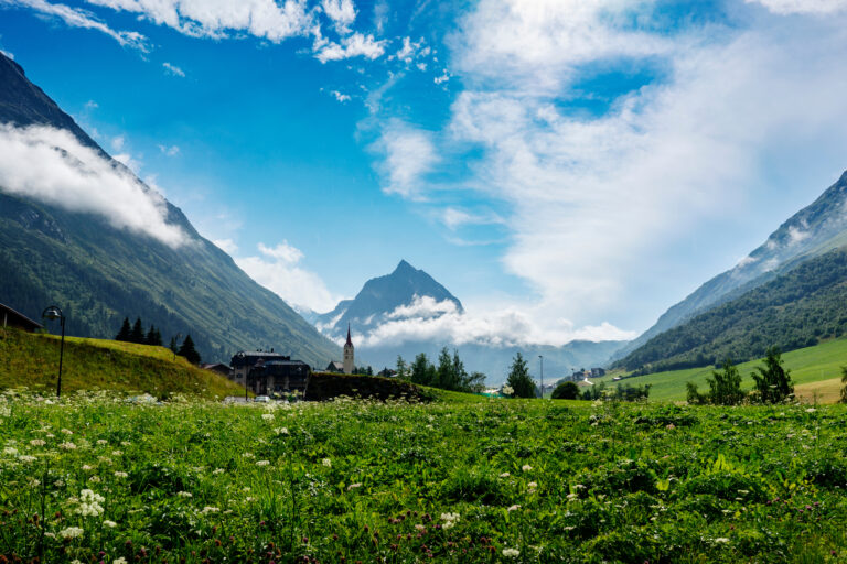 Ein malerischer Blick auf eine üppige grüne Wiese mit weißen Blumen im Vordergrund, die zu einem kleinen Dorf mit einer Kirche führt. Schneebedeckte Berge umgeben das Tal unter einem strahlend blauen Himmel mit vereinzelten Wolken.