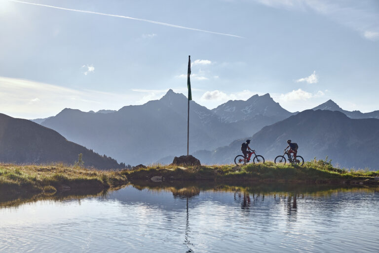 Zwei Radfahrer fahren einen Weg neben einem spiegelnden Bergsee entlang. In der Nähe steht eine Flagge und im Hintergrund ist eine Bergkette unter einem klaren Himmel zu sehen. Die Szene ist ruhig und von sanftem Tageslicht erhellt.